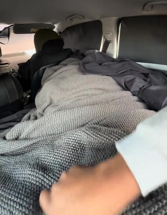 Person sleeping under a grey blanket in a car.
