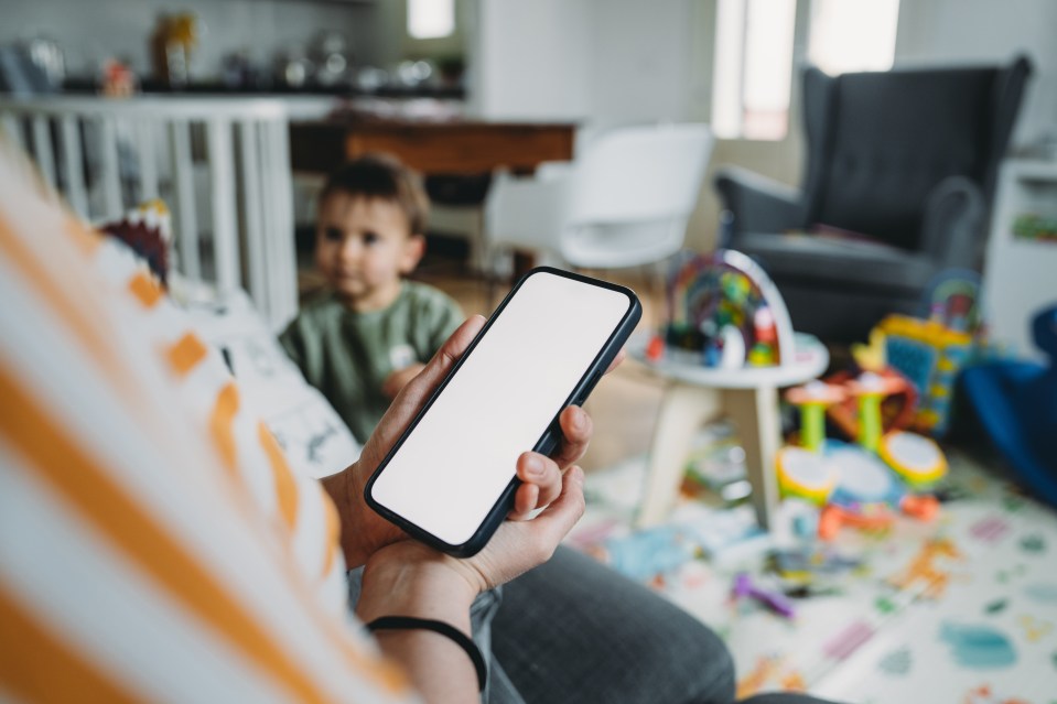 A mother holding a smartphone with a blank screen, with her toddler out of focus in the background.