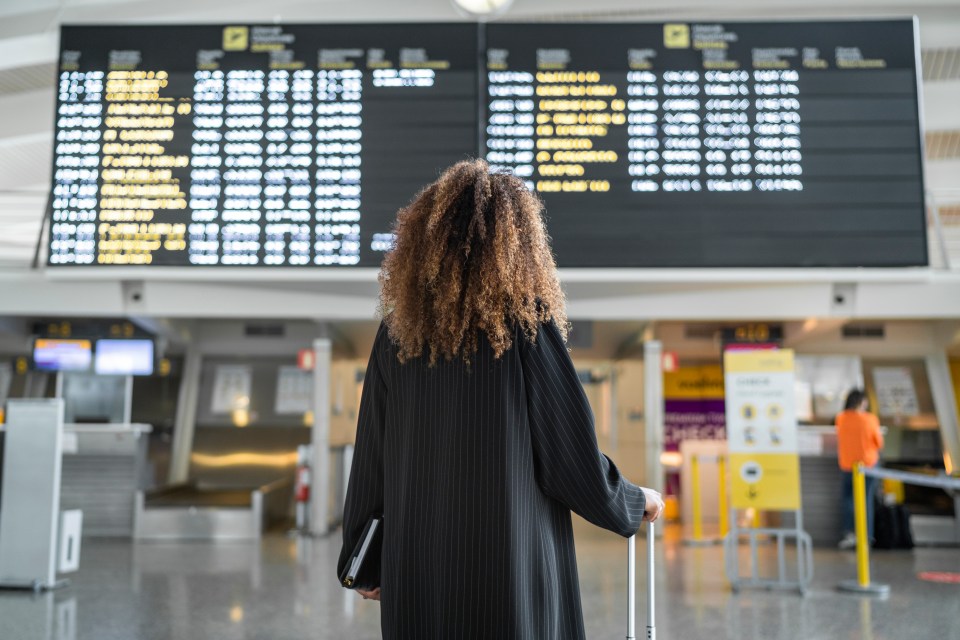 Woman with curly hair at an airport looking at a departures board.