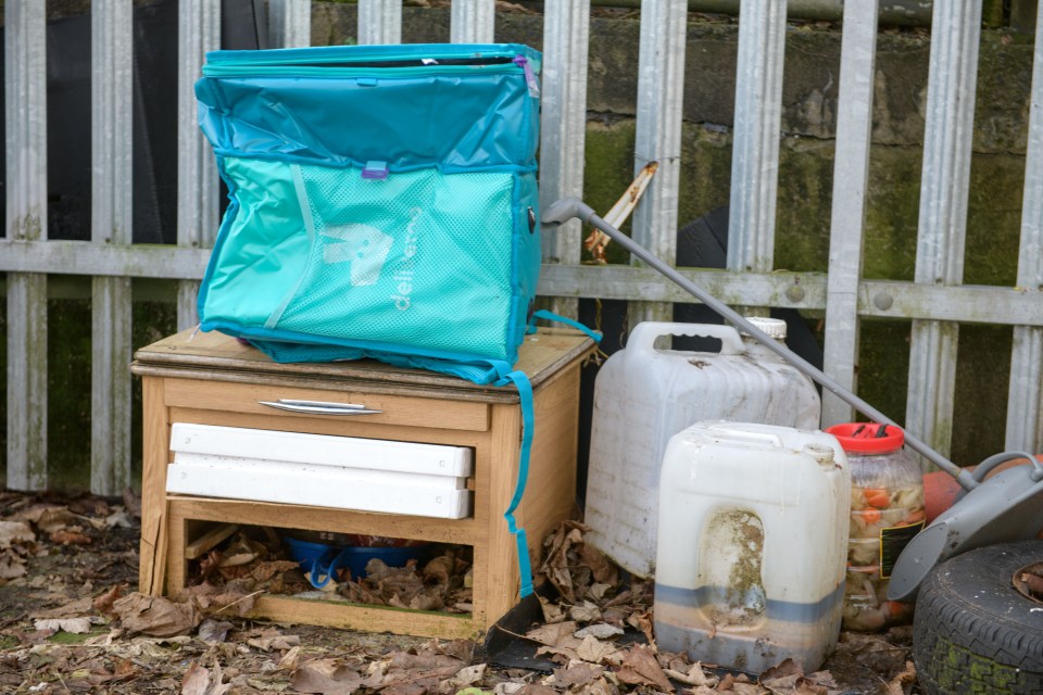 Discarded items including a teal bag, wooden cabinet, and plastic containers near a fence.