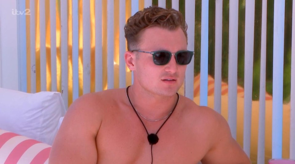 Man wearing sunglasses and necklace, sitting shirtless.