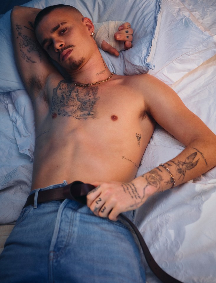 A tattooed man lies on a bed in jeans.