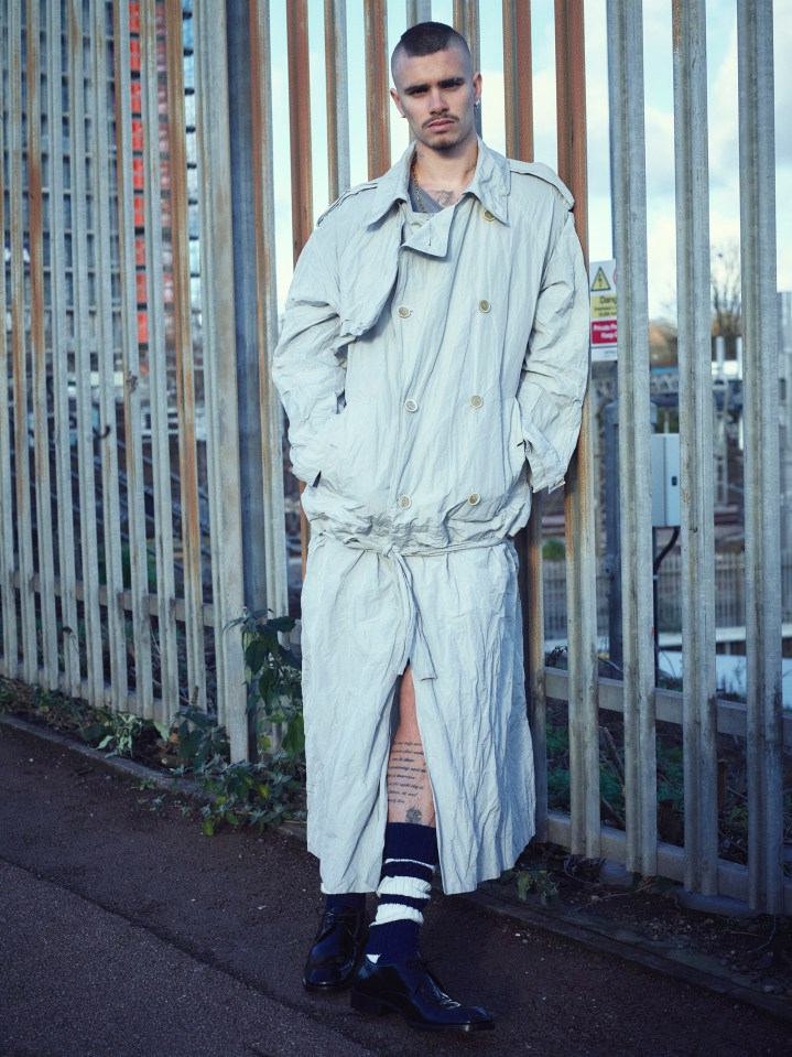 Man in long light gray coat standing by a fence.