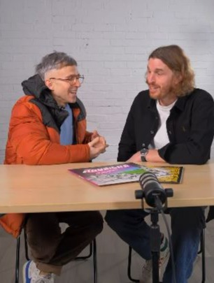 Two men sitting at a table, talking.  A microphone is on the table between them.