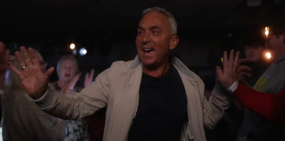 A man in a light beige jacket reacts enthusiastically in a dark room with other people.