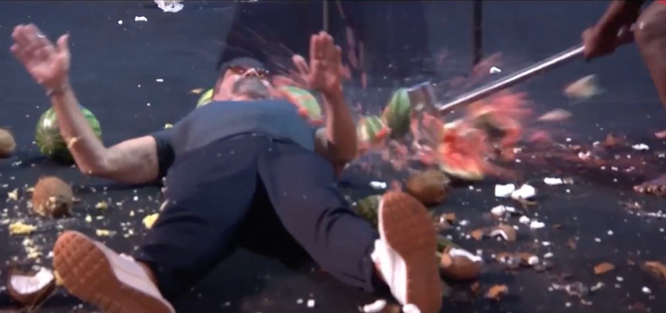Screenshot of a man lying on the ground surrounded by smashed watermelons and coconuts, as someone swings a large blade nearby.