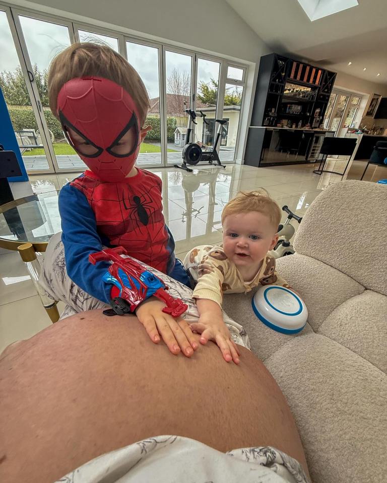 A pregnant woman's large baby bump with two young children touching it; one is wearing a Spider-Man costume.