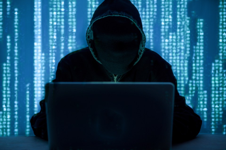 Hooded figure using laptop in front of a screen displaying binary code.