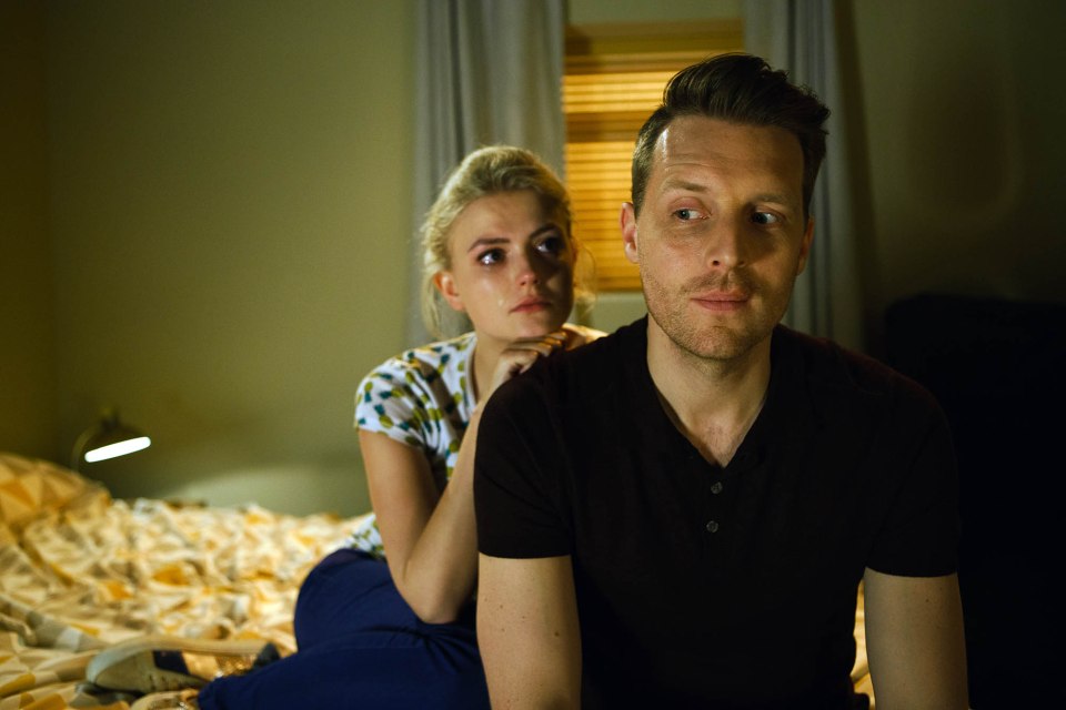 A man and woman sit on a bed, the woman crying and touching the man's shoulder.