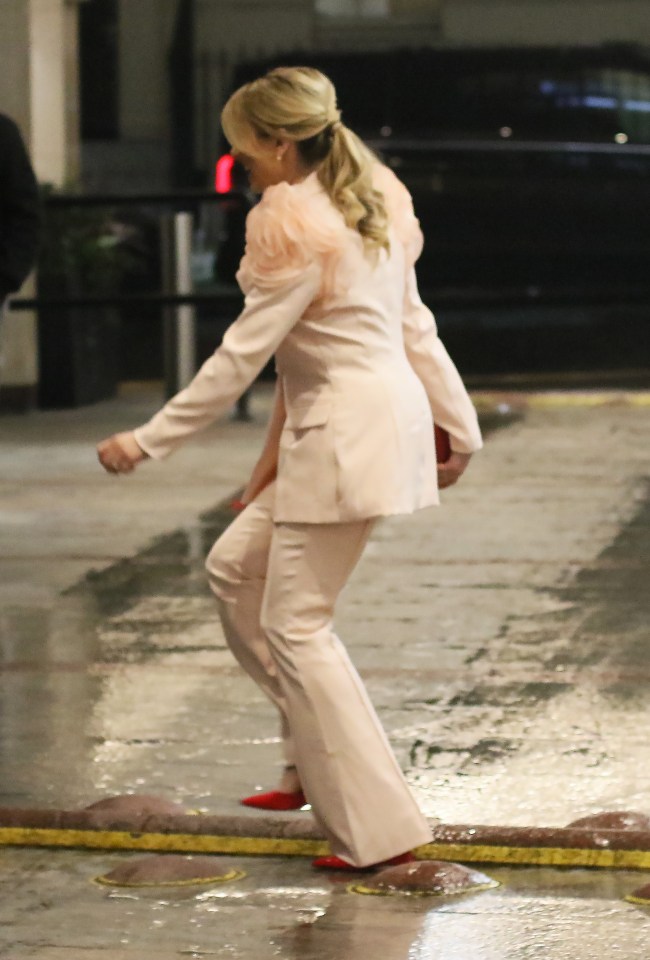 Woman in a light pink pantsuit and red shoes walking on wet cobblestones.