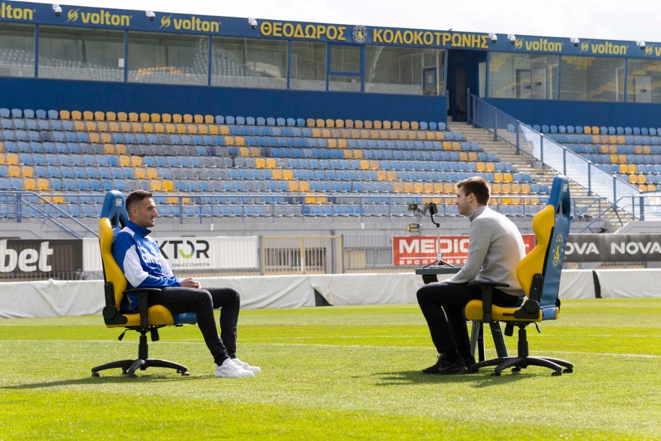 Two men sitting in chairs on a soccer field, facing each other.