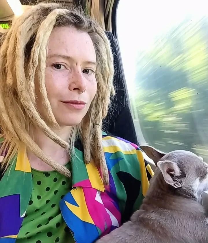 Woman with dreadlocks and colorful shirt holding a small dog on a train.