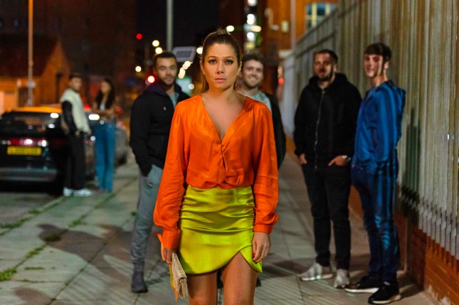 Nikki Sanderson, star of Hollyoaks, walking down a street at night.  A group of men are behind her.