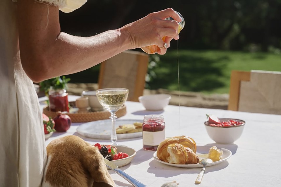 Person pouring honey onto a croissant at an outdoor breakfast.