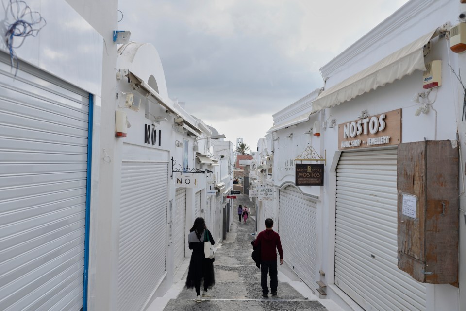A narrow street lined with white buildings and closed shops, two people walking away from the camera.