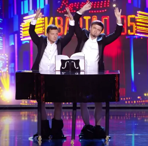 Two men in suits standing behind a grand piano on a stage.