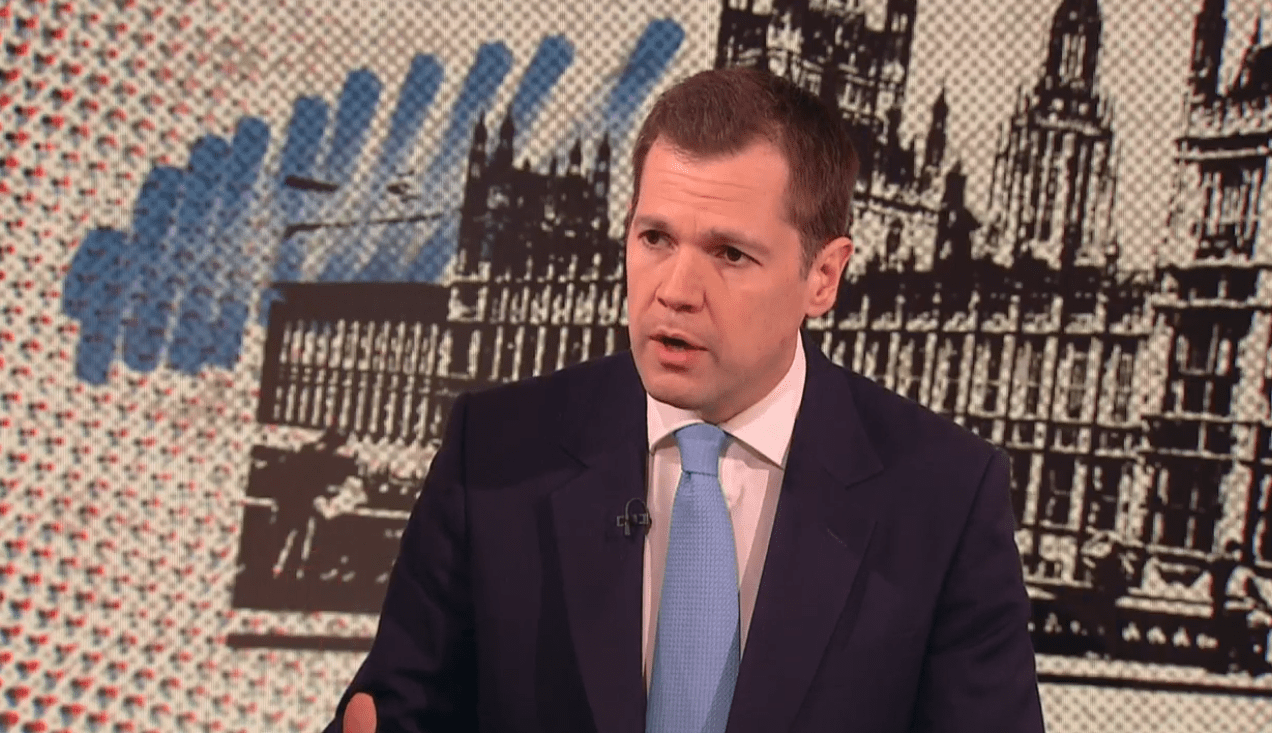 Man in suit speaking in front of a graphic of the Houses of Parliament.
