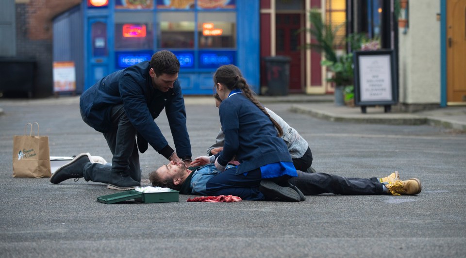 A man lies injured on the ground as two people attend to him.