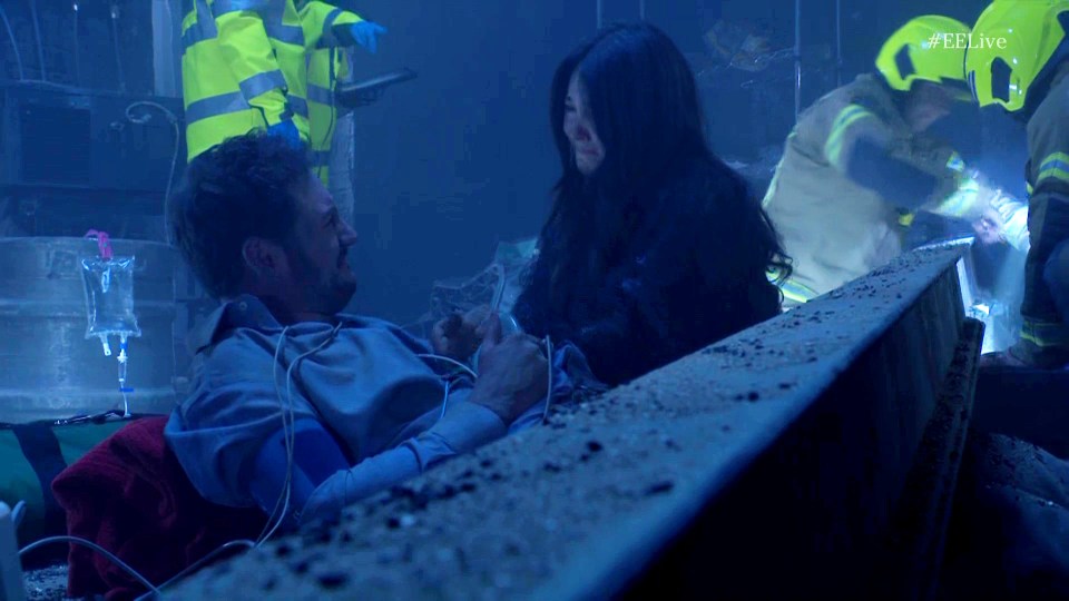 A man lies injured, receiving medical attention, while a woman comforts him.