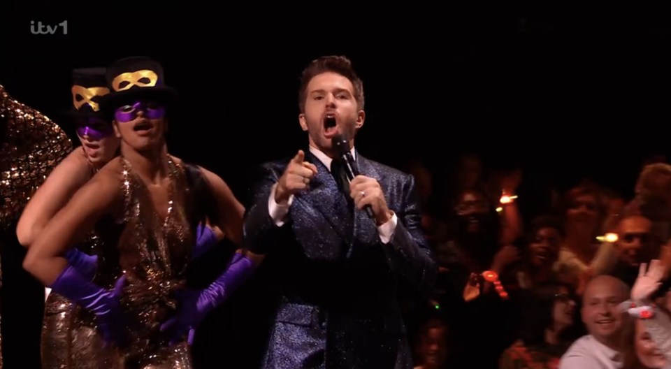 A singer in a sparkly suit performs on stage with two masked dancers.