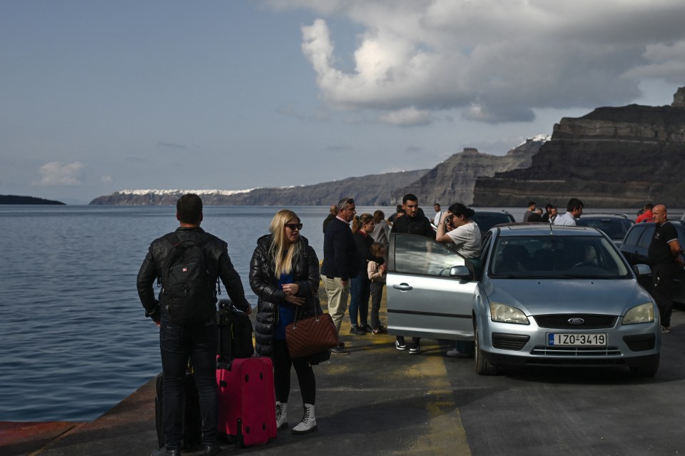 People waiting at a ferry terminal on Santorini, Greece, with luggage and cars.