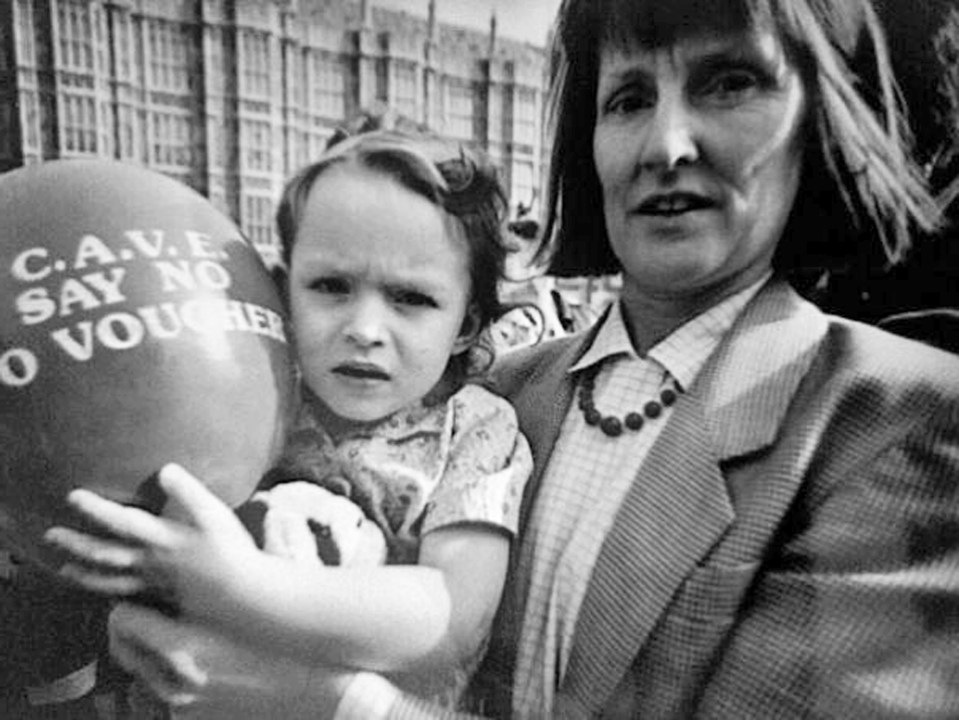 Black and white photo of a woman holding a young child at a protest, holding a balloon that says "C.A.V.E. SAY NO TO VOUCHER".