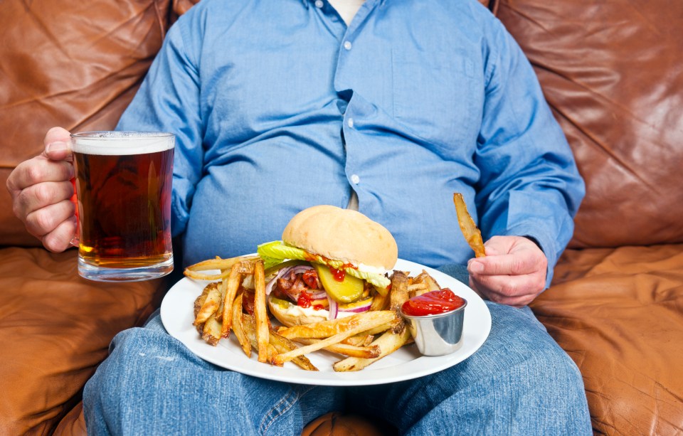 Overweight man sitting on a couch eating a large burger and fries with a beer.
