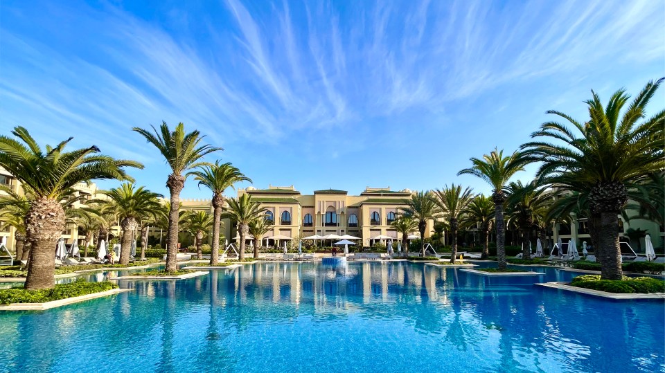 Resort pool with palm trees and building reflected in the water.
