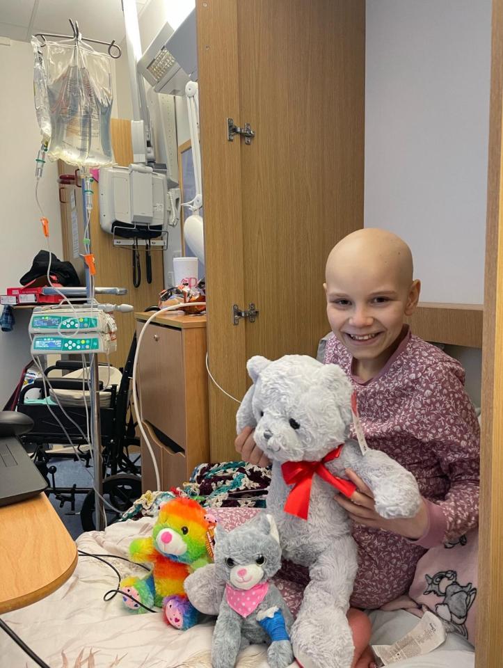 A smiling girl with no hair holds a large stuffed teddy bear in a hospital room.