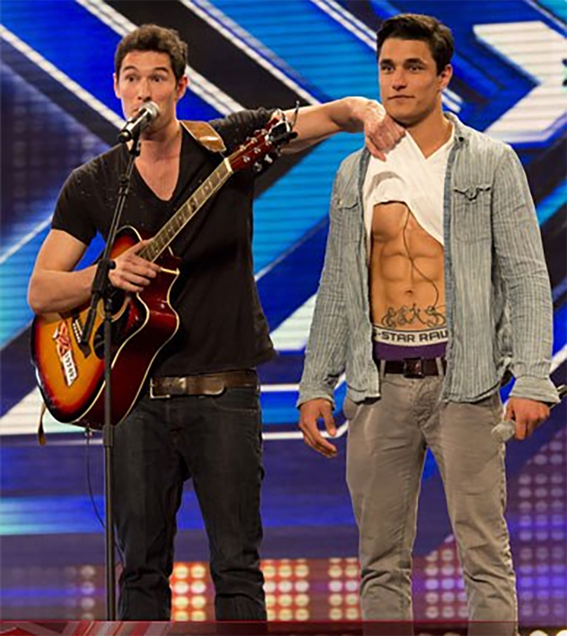 Two men on Britain's Got Talent stage; one plays guitar, the other shows his abs.