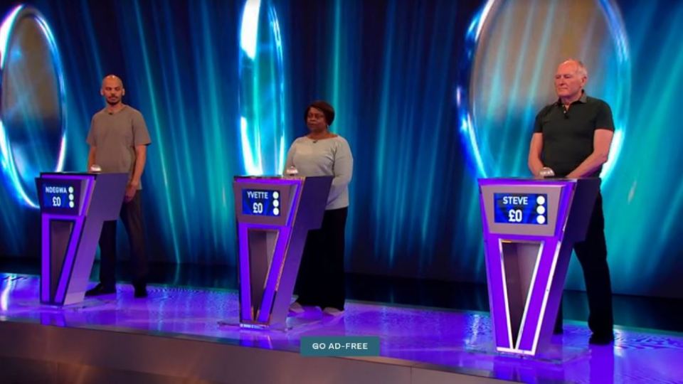 Three contestants stand at podiums on a game show set.