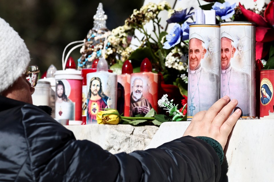A woman's hand rests on votive candles depicting Pope Francis and other religious figures near a statue of Pope John Paul II.