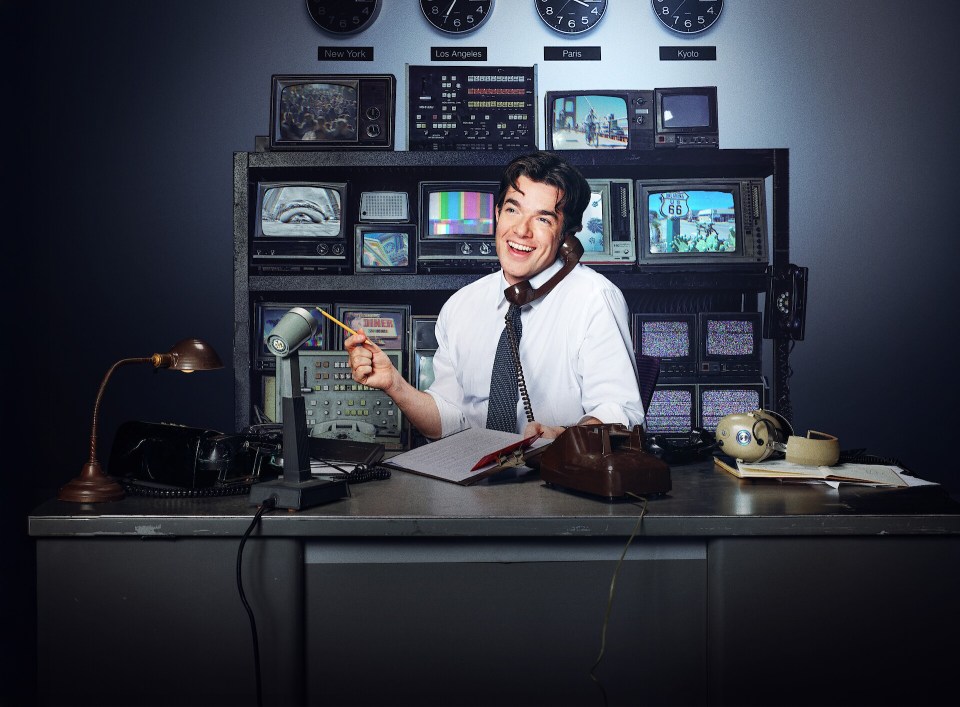 A man sits at a desk in a newsroom, surrounded by vintage television monitors and equipment, speaking on a rotary phone.