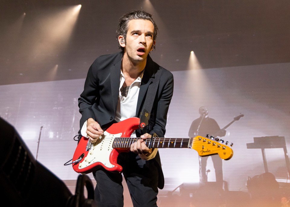Matty Healy of The 1975 playing a red electric guitar on stage.