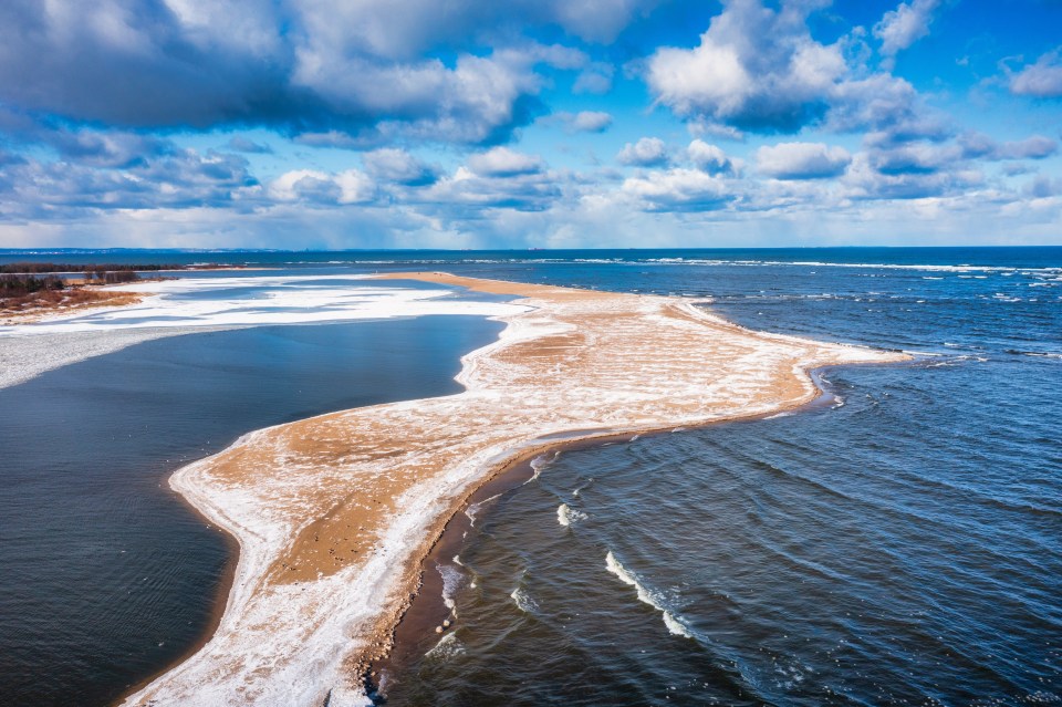 Aerial view of a snow-covered sandbar separating a partially frozen body of water from the ocean.