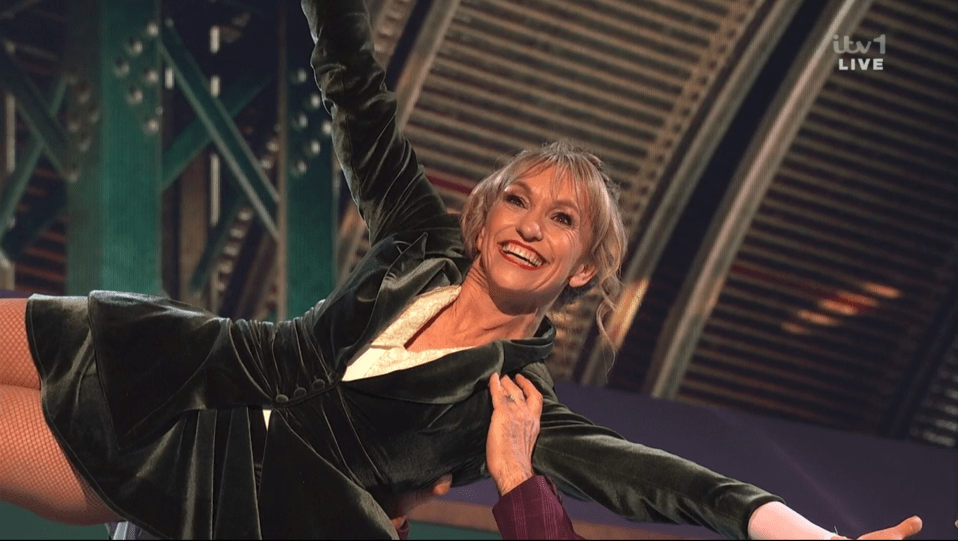 Screenshot of a woman in a green velvet jacket being lifted by a person out of frame.