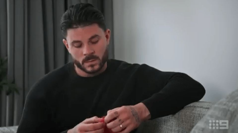 Screenshot of a man sitting on a couch, holding a small red object.