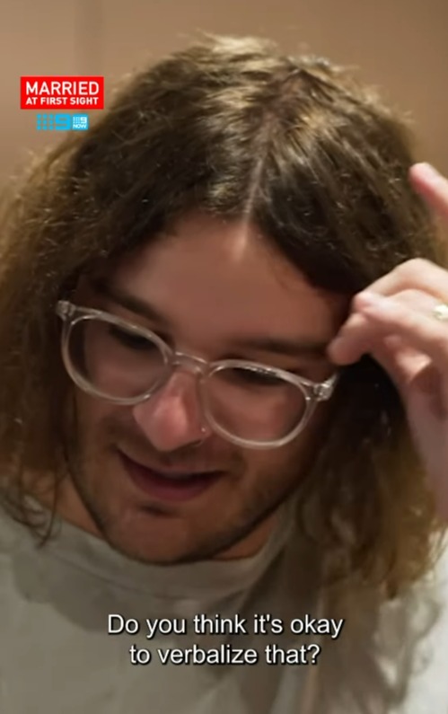 Screenshot of a man with long curly hair and clear glasses, looking down and touching his hair, with the text "Do you think it's okay to verbalize that?" overlaid on the bottom.  The "Married at First Sight" logo is visible in the upper left corner.