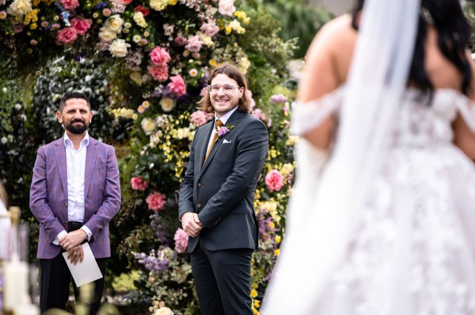 Ashleigh and Jake's wedding. Groom stands before a floral wall.  A guest in a purple jacket and the bride's back are visible.