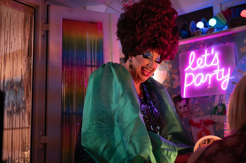 Mark Benton as Mam in a sparkly outfit and large red wig, smiling in front of a "Let's Party" neon sign.
