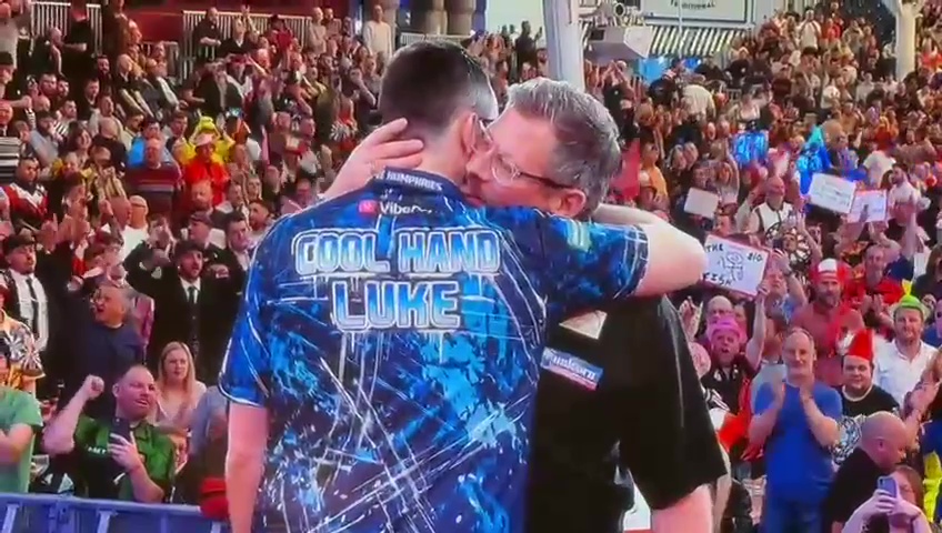 James Wade kissing Luke Humphries on the neck at the UK Open, surrounded by spectators.