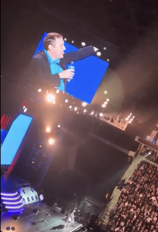 Peter Kay on a large screen at a concert, gesturing.