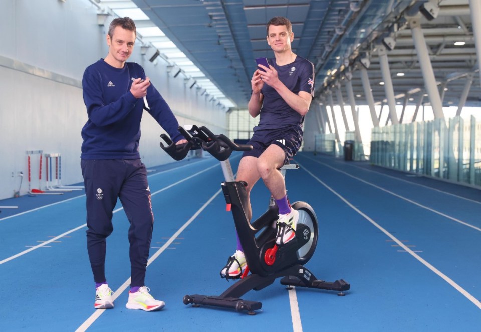 Two Olympic athletes on an indoor track, one sitting on a stationary bike, the other standing beside him.