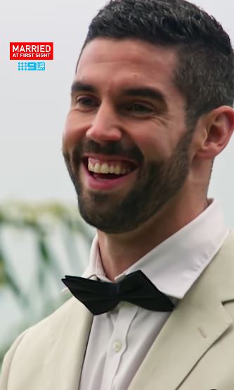 A smiling groom in a beige suit and bow tie at his wedding.