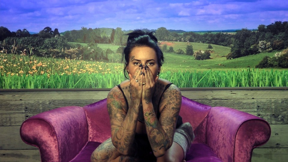 A heavily tattooed woman sits on a purple armchair, hands covering her face; a pastoral scene is visible in the background.
