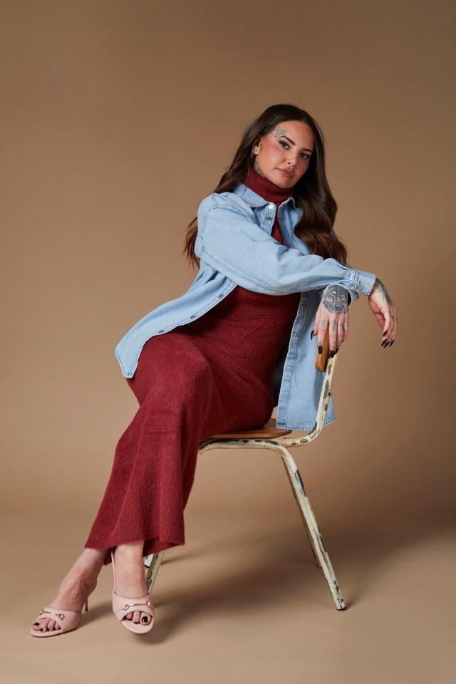 Jemma Lucy in a maroon dress and denim shirt, sitting on a chair.