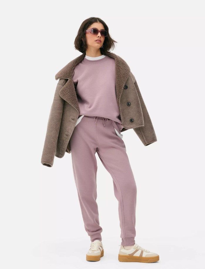 Woman wearing mauve joggers and sweatshirt with a brown jacket.