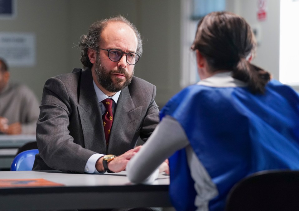 A man in a suit sits across a table from a woman in a blue jumpsuit, appearing to be in a prison interview.