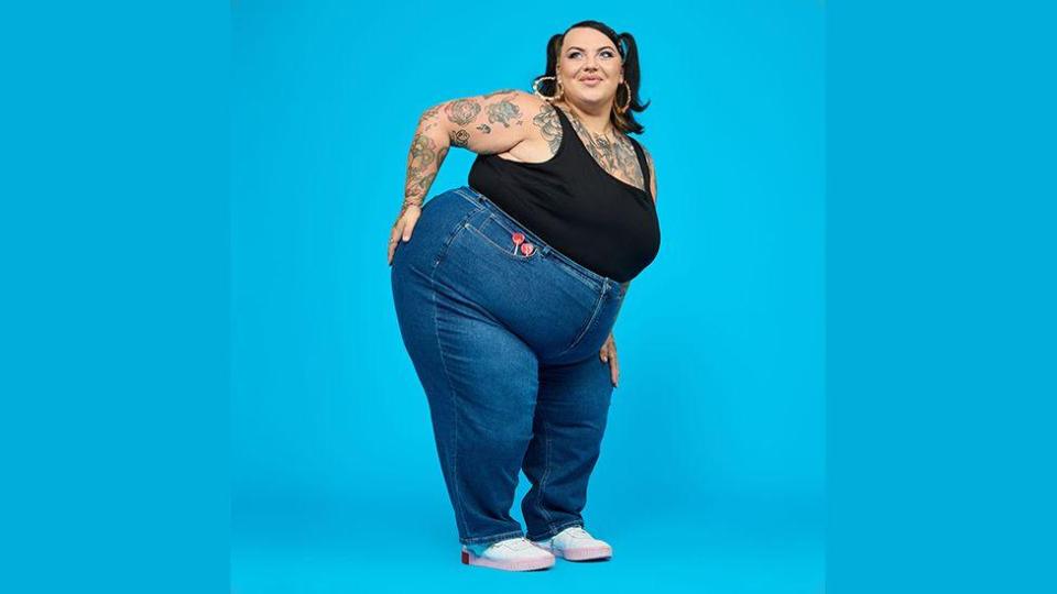 Plus-size model wearing jeans and a black tank top against a blue background.