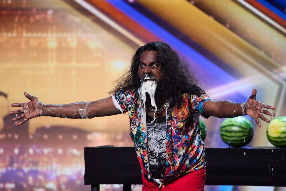 A man with long hair and a colorful shirt sprays milk from his mouth during a performance.